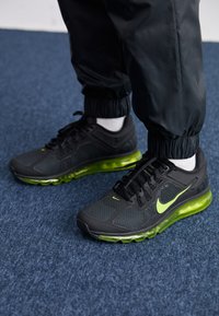 Sapatos de desporto negros com tecido em malha, sola translúcida verde e detalhes amarelos. Apresentam atacadores e uma palmilha almofadada para suporte.