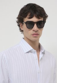 Jeune homme aux cheveux noirs et bouclés, portant des lunettes de soleil noires et une chemise blanche à rayures, sur un fond clair uni.