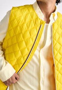 Uomo che indossa una camicia di un giallo chiaro e sta aprendo un gilet squadrato di un giallo brillante con zip a righe.