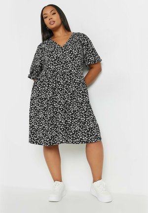 Schwarzes Blumenkleid mit V-Ausschnitt, kurzen Ärmeln und ausgestellt geschnittenem Saum. Mit weißen Blumen durchzogen und kombiniert mit weißen Sneakern.
