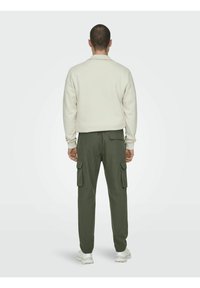 Helles beiges Sweatshirt mit Stehkragen, kombiniert mit olivgrünen Cargo-Hosen mit seitlichen Taschen. Das Material wirkt weich und lässig.