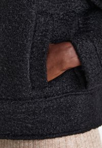 Poche d'un manteau gris foncé texturé avec une main à la peau foncée partiellement à l'intérieur, et un tissu côtelé beige visible sous le manteau.