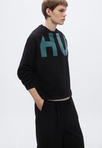 Μαύρο φούτερ με μεγάλο γραφικό «HU» σε τιρκουάζ απόχρωση στο μπροστινό μέρος, με ριμπ μανσέτες, άνετη εφαρμογή, συνδυασμένο με μαύρο παντελόνι, το μοντέλο στέκεται πλευρικά.