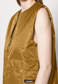 Light brown sleeveless vest with a high neckline, zippered front, two large front pockets, and a black label at the hem. Smooth, shiny fabric.