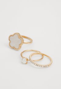 Set di tre anelli d'oro: uno presenta una forma di fiore bianco con un bordo testurizzato, un altro ha un dettaglio di perla e il terzo è decorato con piccole pietre trasparenti.