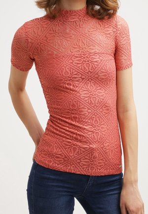 Femme portant un haut corseté en dentelle corail à manches courtes avec des motifs géométriques et floraux, associé à un jean bleu foncé.