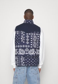 Blå vadderad väst med vita paisley- och geometriska mönster, med en ribbad krage och en kontrasterande överdel. Texturerad och visuellt slående.