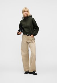 Γυναίκα με σκουρό πράσινο φούτερ με κουκούλα, μπεζ παντελόνι φαρδιάς γραμμής και μαύρα παπούτσια, κρατώντας μια μαύρη τσάντα και στέκεται μπροστά σε απλό φόντο.