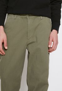 Pantalon en coton kaki avec une taille élastique, des poches latérales et une coupe décontract ée, présenté avec des mains reposant sur le tissu.