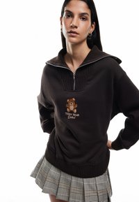 Brązowy sweatshirt z ribbonowym kołnierzem i zamkiem błyskawicznym 1/4; ozdobiony haftowanym wzorem misia z napisem "Teddy Bear District." Spódnica plisowana w kratkę.
