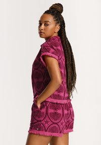 Žena s dlouhými spletenými vlasy, oblečená v fialovém vzorovaném tričku s krátkým rukávem a odpovídajících šortkách, s rukama v kapsách a dívající se přes rameno.