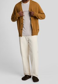 Cárdigan marrón con grandes botones, usado sobre una camisa gris clara, combinado con pantalones blanco roto y zapatos marrón de tipo slip-on. Textura suave.