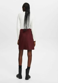 Jupe bordeaux à coupe droite, texture laineuse, portée avec des bottines noires. Le modèle a de longs cheveux tressés et un haut blanc à côtes.