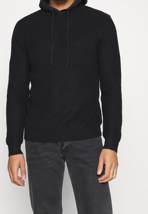 Suéter negro con textura y capucha, con un patrón tejido, puños y dobladillo acanalados. Combinado con jeans gris oscuro.