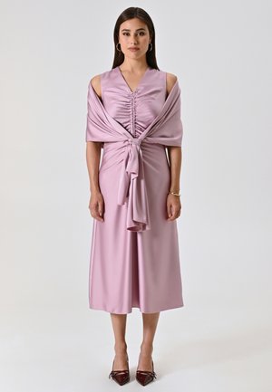 Abito in satin lavanda con arricciature all'polso, maniche a spalle scoperte e dettaglio con fiocco in vita; abbinato a tacchi bordeaux.