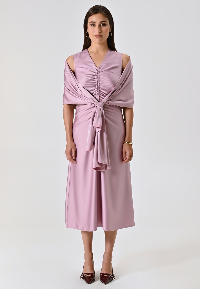 Abito in satin lavanda con arricciature all'polso, maniche a spalle scoperte e dettaglio con fiocco in vita; abbinato a tacchi bordeaux.