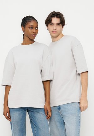 Doi tineri adulți, o femeie și un bărbat, purtând tricouri albe supradimensionate și simple, precum și blugi albaștri, stau unul lângă altul pe un fundal alb.