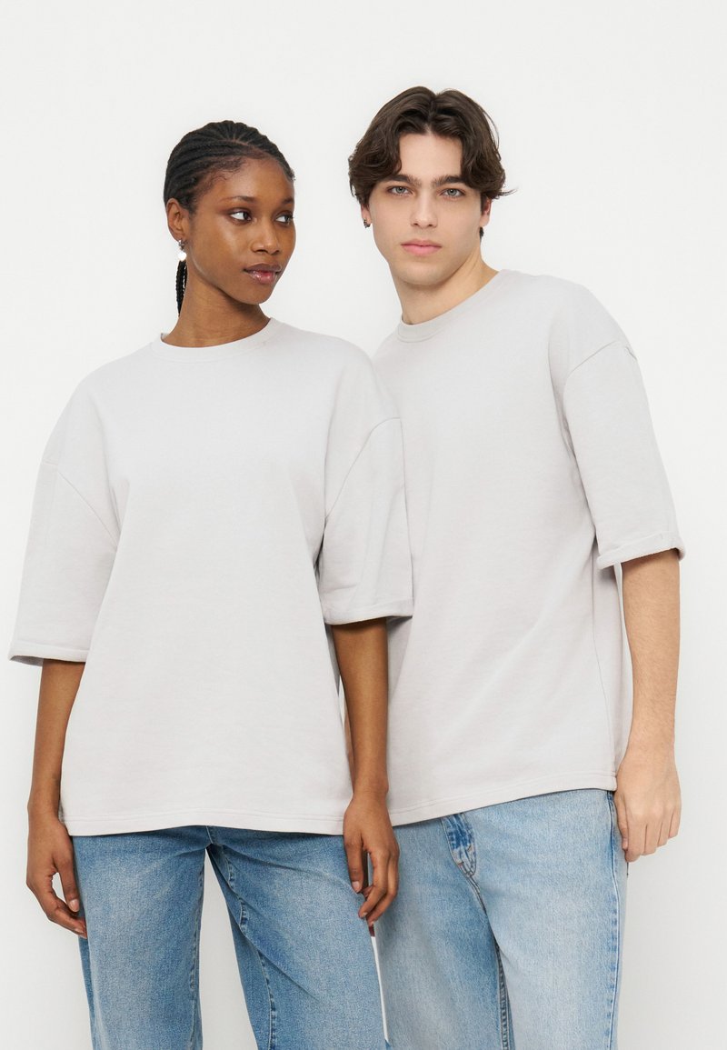 Δύο νεαροί ενήλικες, ένας άνδρας και μία γυναίκα, φορούν απλά, φαρδιά λευκά μπλουζάκια και μπλε τζιν, στέκονται δίπλα δίπλα μπροστά σε λευκό φόντο.