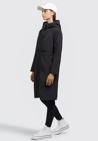 Veste longue noire coupe-vent avec capuche, dotée d'une fermeture éclair sur le devant, de poches latérales et d'une coupe ample. Associée à des leggings noirs et des baskets blanches.