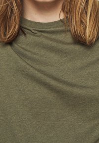 T-shirt a girocollo verde oliva realizzato in tessuto morbido e testurizzato, con leggere pieghe e cuciture lisce, primo piano sui dettagli del tessuto.