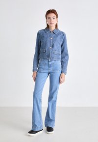 Veste en denim bleu coupée avec un col, des poches avant et des boutons dorés. Associée à un jean flare bleu clair et des baskets noires matelassées.
