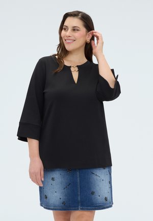 Žena v černém topu se zlatou sponou a tříčtvrtečními rukávy, doplněném modrou džínovou sukní zdobenou malými květinovými vzory.