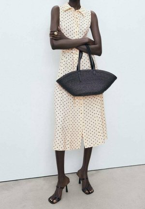 Leichtgelbes Midi-Kleid mit Polka-Dots und Kragen, kombiniert mit einer schwarzen geflochtenen Tragetasche und schwarzen Sandalen mit Absatz und Riemchen.