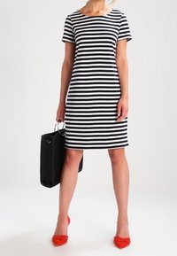 Kvinna som bär en svartvit randig klänning med horisontella ränder, håller en svart handväska och har på sig klarröda spetsiga klackskor.