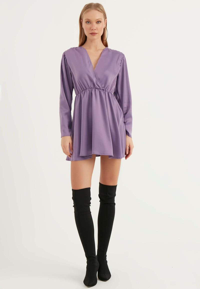 Lila langärmliges Kleid mit V-Ausschnitt und tailliertem Bund; der fließende Rock reicht bis zur Mitte des Oberschenkels. Das Model trägt schwarze Overknee-Stiefel.