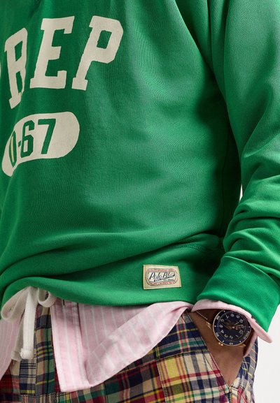 Zaļš kokvilnas džemperis ar baltu uzrakstu "REP 0-67", uzvilkts virs rozā svītrota krekla un pjūtas šortiem, ar Polo Ralph Lauren logotipa uzlīmi.