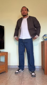 Grå bomberjakke med ribbestrikkede mansjetter over en hvit t-skjorte, kombinert med blå jeans og svart-hvite joggesko. Stående innendørs.