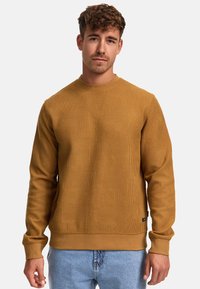 Sudadera de color mostaza con un patrón texturizado, cuello redondo y puños de canalé. Combinada con unos jeans de mezclilla claros, mostrando un aspecto casual.