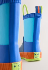 Bottes de pluie en caoutchouc bleu vif et orange avec semelles vertes et boucles d'attache, ornées d'un visage souriant jaune qui fait un clin d'œil sur les talons.