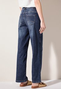 Jeans en denim bleu foncé taille haute avec un design à jambes droites. Dotés de poches arrière et d'une texture subtile. Portés avec des sandales tressées marron.