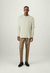 Off-white Kabelstrickpullover, beiger Hose und schwarze Sneaker mit Gummisohlen. Das Outfit hat eine lockere Passform und ein strukturiertes Design.