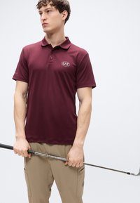 Μπορντό πόλο μπλούζα με γιακά και κοντά μανίκια, με οβάλ λογότυπο EA7 στο στήθος, συνδυασμένη με χακί παντελόνι και ένα μπαστούνι γκολφ.