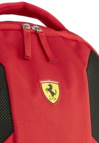 Zaino in tessuto rosso con dettagli in rete nera, caratterizzato da un logo Ferrari giallo e nero in evidenza, doppie zip e una maniglia imbottita.