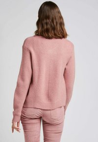 Pull en tricot rose clair avec une texture côtelée, des manches tombantes et une coupe décontractée. Le dos présente un détail de couture le long de la colonne vertébrale.