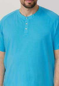 Błękitna koszula krótki rękaw z okrągłym dekoltem i zakładką na cztery guziki. Miękki, gładki materiał o luźnym kroju i rękawach raglanowych.