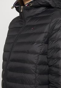 Zwarte donzen jas met doorgestikt ontwerp, voorzien van een hoge kraag, ritssluiting en logo-accent. Gladde, glanzende textuur met capuchon.