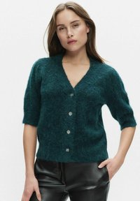 Sarviininsininen, pehmeä neulottu neuletakki, jossa on v-kaula-aukko, lyhyet hihat ja viisi hopeista nappia edessä. Päällä mustat keinonahkahousut.