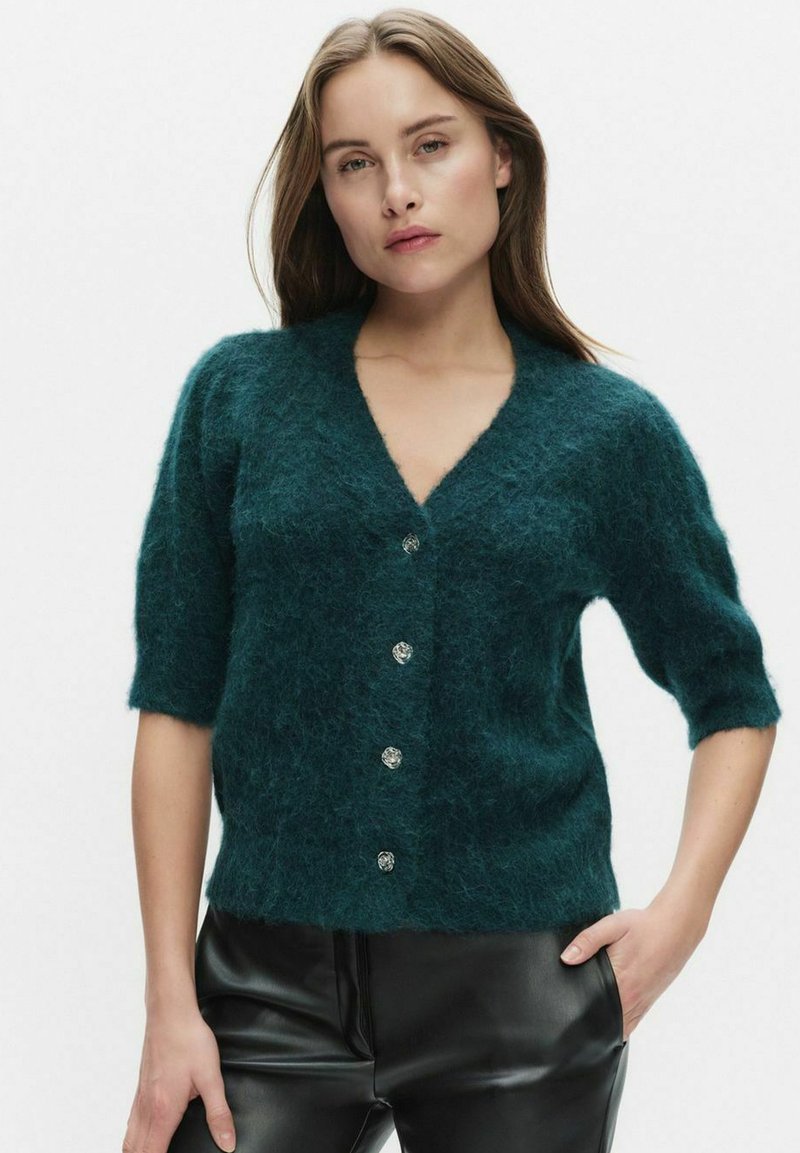 Sarviininsininen, pehmeä neulottu neuletakki, jossa on v-kaula-aukko, lyhyet hihat ja viisi hopeista nappia edessä. Päällä mustat keinonahkahousut.