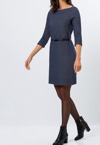 Modrá pletená šatová šaty s tříčtvrtečními rukávy, páskem v pase a délku ke kolenům. Spárováno s černými kotníkovými botami a průhlednými punčochami.
