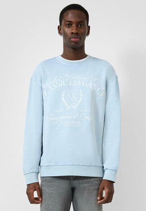 Jonge man die een lichtblauwe sweatshirt draagt met witte tekst en afbeeldingen, staand tegen een effen grijze achtergrond.