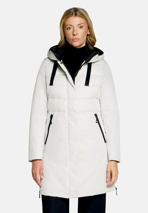 Vrouw draagt een lange witte winterjas met capuchon, zwarte ritstassen en zwarte trekkoorden over een zwart topje.