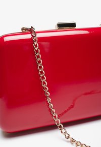 Pochette en cuir verni rouge avec une chaîne en doré et un fermoir doré. Surface lisse et brillante avec une forme rectangulaire.