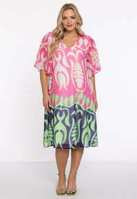 Robe à col en V, à manches courtes, de couleur rose avec un motif de feuilles vertes et bleu foncé. Tissu léger et fluide. Assortie avec des sandales à lanières vertes.