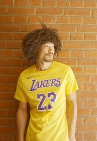 Gul bomullst-skjorte med "LAKERS" og nummeret "23" i lilla. Korte ermer, avslappet passform, står mot en murvegg.