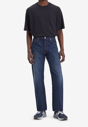 Personne portant un t-shirt à poches bleu marine foncé, un jean bleu coupe droite et des baskets bleu marine et grises, debout devant un fond blanc uni.