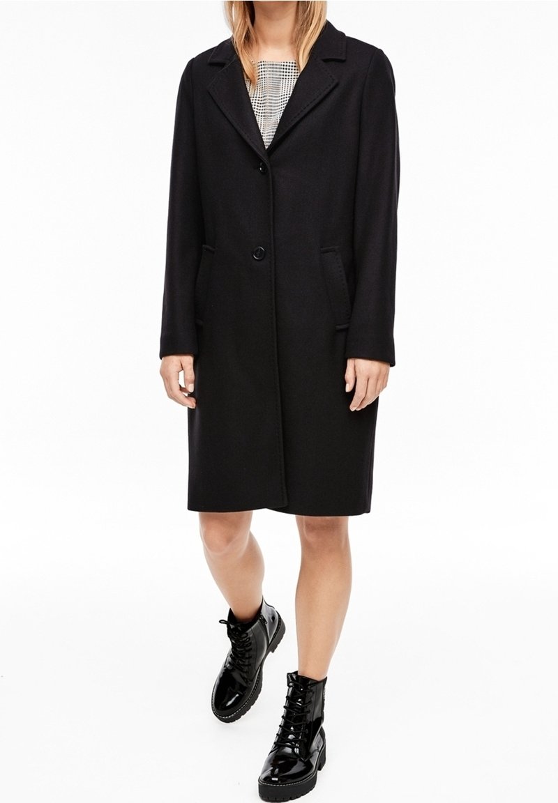 Femme portant un manteau noir mi-longueur avec deux boutons et des poches, bottines noires brillantes à lacets, debout sur fond blanc.